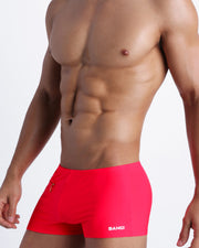 Left side view of a masculine model wearing men’s swimwear in a red color with official logo of BANG! Brand in white.
