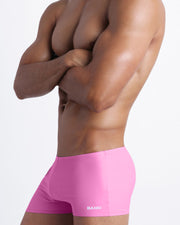 Left side view of a masculine model wearing men’s compression swimwear shorts in bubble gum pink color featuring a side pocket with official logo of BANG! Brand.