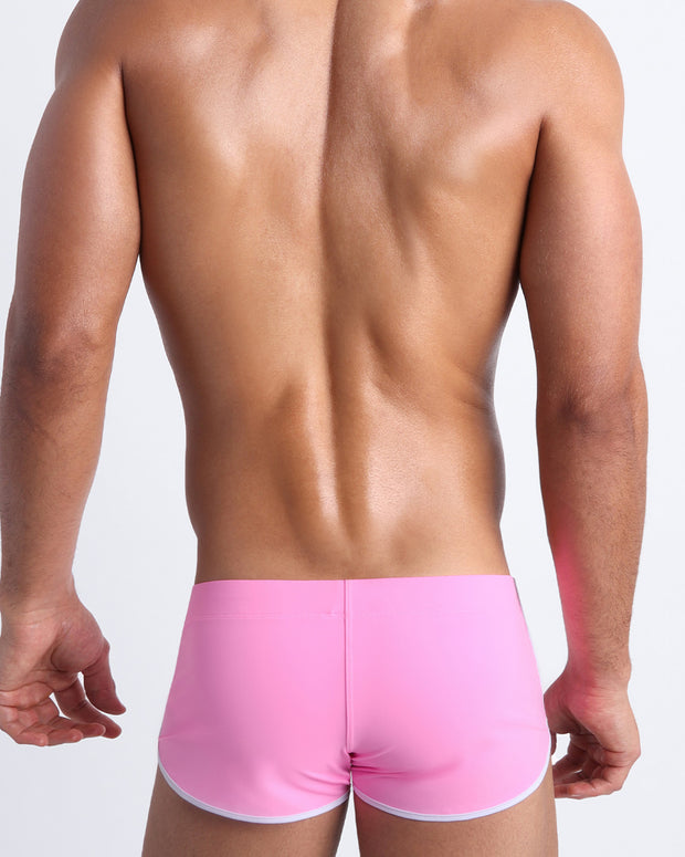 Back view of a male model wearing men’s swim mini shorts in bubble gum pink by the Bang! Clothes brand of men&