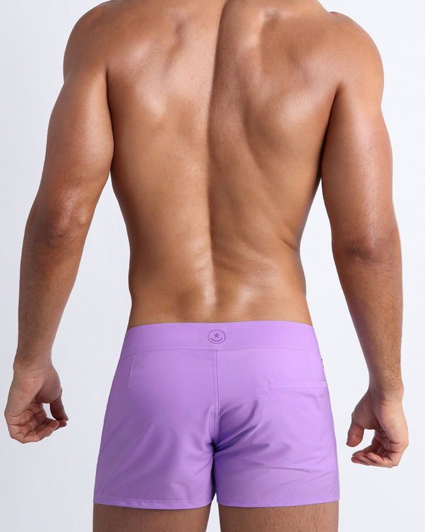 Back view of a male model wearing men’s beach trunks in neon light purple color by the Bang! Clothes brand of men&