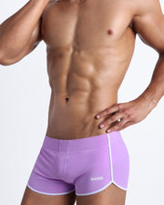 Left side view of a masculine model wearing men’s swimwear beach shorts in lavender color made with Italian-made Vita By Carvico Econyl Nylon with official logo of BANG! Brand in white.