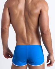 Back view of a male model wearing men’s swim shorts in blue by the Bang! Clothes brand of men's beachwear from Miami.