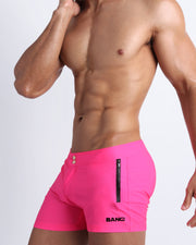 Left side view of a masculine model wearing men’s swim shorts in neon pink color featuring a side pocket with official logo of BANG! Brand.