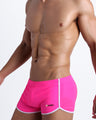 Left side view of a masculine model wearing men’s swimwear in neon pink with official logo of BANG! Brand in white.