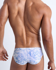 Back view of male model wearing the SPLASH  beach briefs for men by BANG! Miami in white and blue color with graphics of water and wet letters.