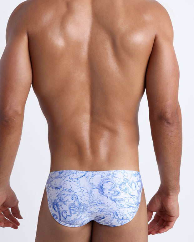 Back view of male model wearing the SPLASH  beach mini-briefs for men by BANG! Miami in white and blue color with graphics of water and wet letters.