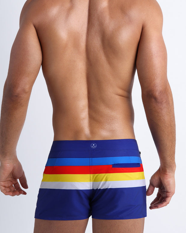 Back view of male model wearing  swim beach shorts in navy blue color with color stripes in white, yellow, bold red, and blue.