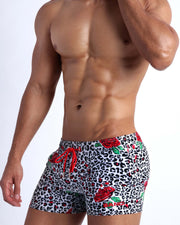 Lateral view of a man wearing the SO RED THE ROSE shorter leg length swim trunks by Bang! Clothes black & white leopard animal print with red roses.