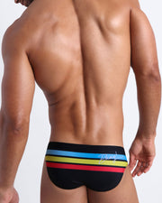 Back side view of male model wearing men's swimwear in black color with color stripes in turquoise blue, yellow, and red.