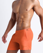 Left side view of a masculine model wearing men’s swimsuit shorts in apricot color featuring a side pocket with official logo of BANG! Brand.
