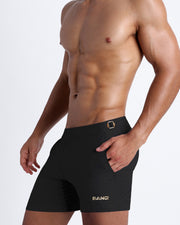 Left side view of a masculine model wearing men’s swimsuit shorts in black featuring a side pocket with official logo of BANG! Brand in gold.