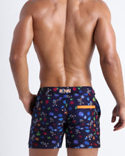 Back view of model wearing the HEY MISTER TJ (CLUB MIX) Men’s beach trunks by BANG! with clubbing and disc-jockey details in dark colors.