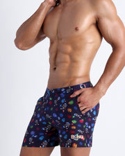 Side view of model wearing the HEY MISTER TJ (CLUB MIX) Men’s shorts for the beach by Bang! Miami, with dark blue background.