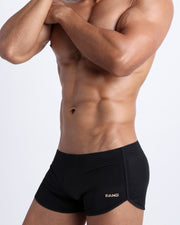 Left side view of a masculine model wearing men’s swimsuit in solid black with official logo of BANG! Brand in gold.