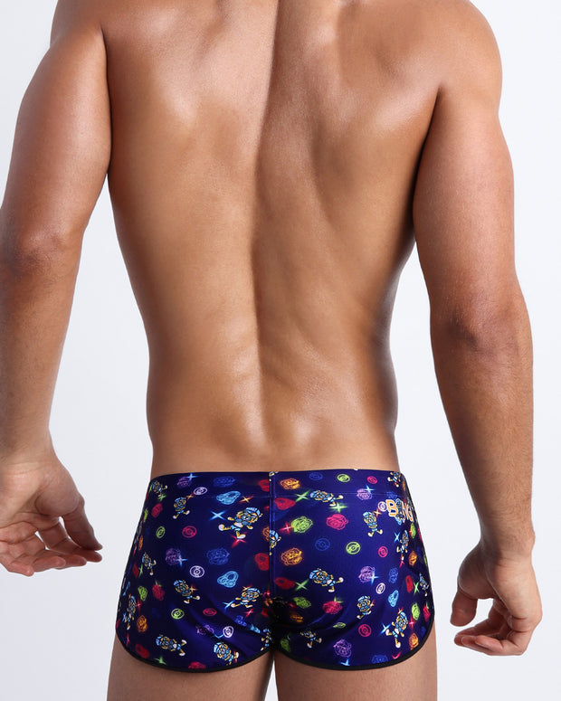 Back view of model wearing the HEY MISTER TJ (CLUB MIX) Men’s beach swim shorts by BANG! with clubbing and disc-jockey details in dark colors.