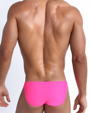 Back view of a male model wearing men’s swim mini-brief in bright pink color by the Bang! Clothes brand of men's beachwear.