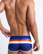 Back view of male model wearing a swim shorts in blue color with color stripes in white, yellow, bold red, and blue.
