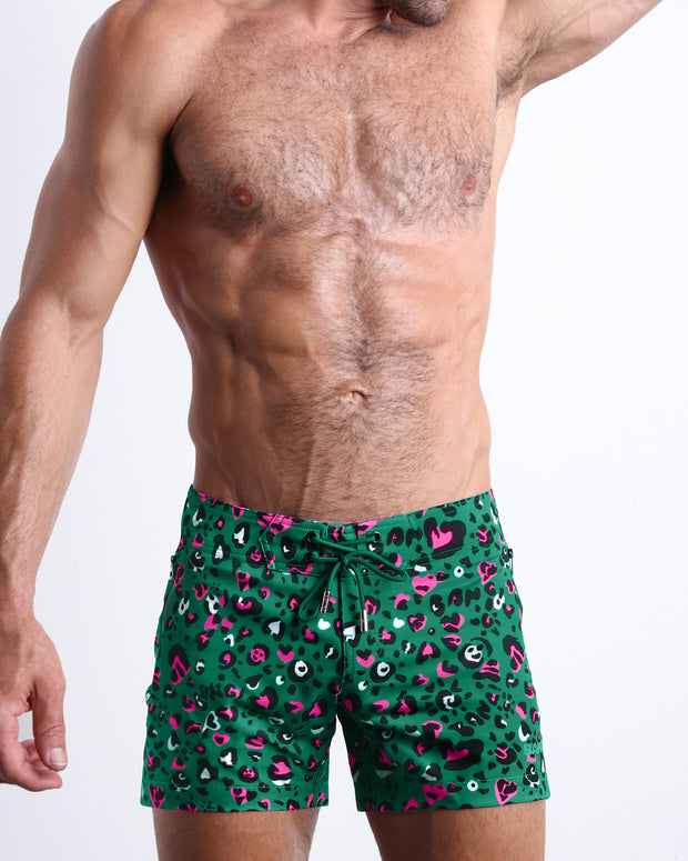 Frontal view of model wearing the CAMO CHAMELEON men’s mini swimming shorts in a forest green color with white and pink camo print by the Bang! menswear brand. 	