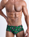 Frontal view of a sexy male model wearing men’s Brazilian Sunga swimsuit in a forest green color with white and pink camo print by the Bang! Menswear brand from Miami.