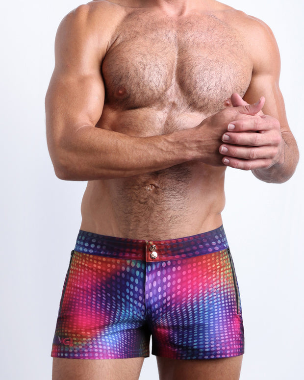Front view of model wearing the CONFESSIONS ON A SAND FLOOR men’swim bottoms inspired by the 80s disco ball music scene in a blue, purple, yellow, green pop of color by the Bang! Clothes brand of men&