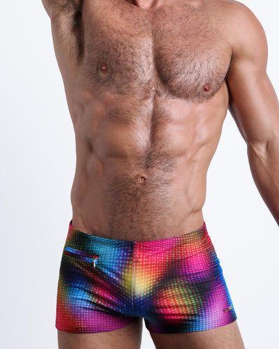 Frontal view of a sexy male model wearing men's swimsuit with mini pockets in CONFESSIONS ON A SAND FLOOR in a pop color disco ball print in purple, blue, red and yellow designed by the Bang! Menswear brand from Miami.