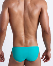 Back view of a male model wearing men’s swim mini-brief in azure color by the Bang! Clothes brand of men's beachwear.