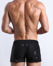 Back view of a male model wearing DARK KNIGHT men’s swim mini shorts with a back zipper pocket in a black color by the Bang! Clothes brand of men's beachwear.
