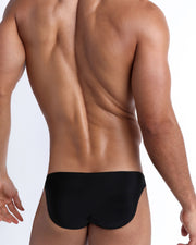 Back view of a male model wearing men’s swim mini-brief in black by the Bang! Clothes brand of men's beachwear.