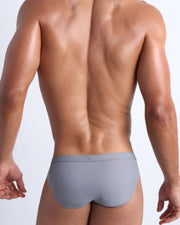 Back view of a male model wearing men’s swim briefs in stone color by the Bang! Clothes brand of men's beachwear.