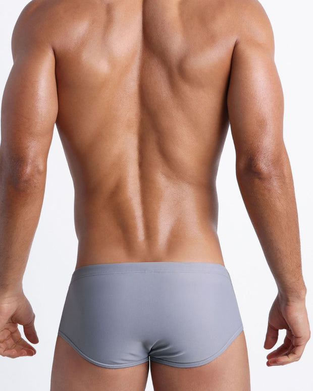 Back view of a male model wearing men’s swim sungas in stone grey color by the Bang! Clothes brand of men&