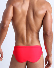 Back view of a male model wearing men’s swim briefs in neon red by the Bang! Clothes brand of men's beachwear.
