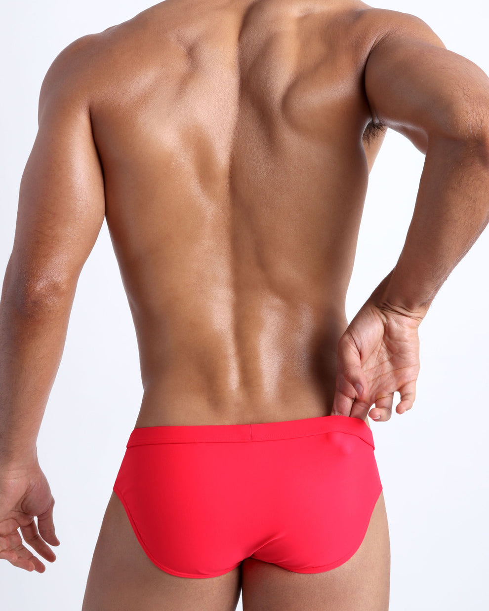 Back view of a male model wearing men’s swim briefs in neon red by the Bang! Clothes brand of men's beachwear.
