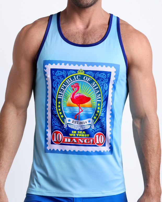Front view of model wearing the IN SUN WE TRUST men’s beach tank top in a light sky blue color and a Miami flamingo pop art postage stamp on the chest by the Bang! Clothes brand of men&
