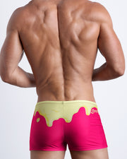 Back view of a Male model wearing swim trunks for men in magenta pink and a pale yellow melting ice cream print by the Bang! Clothes brand of men's beachwear.