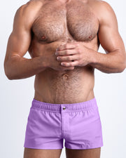 Frontal view of a sexy male model wearing the NEO VIOLET men’s square leg swim trunks in a solid lilac color by the Bang! Menswear brand from Miami.