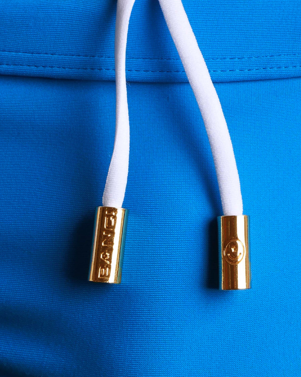 Close-up view of men’s summer swimwear by BANG! clothing brand, showing white cord with custom branded golden cord ends, and matching custom eyelet trims in gold.