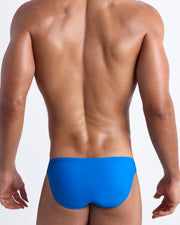 Back view of a male model wearing men’s swim mini-brief in blue by the Bang! Clothes brand of men's beachwear from Miami.