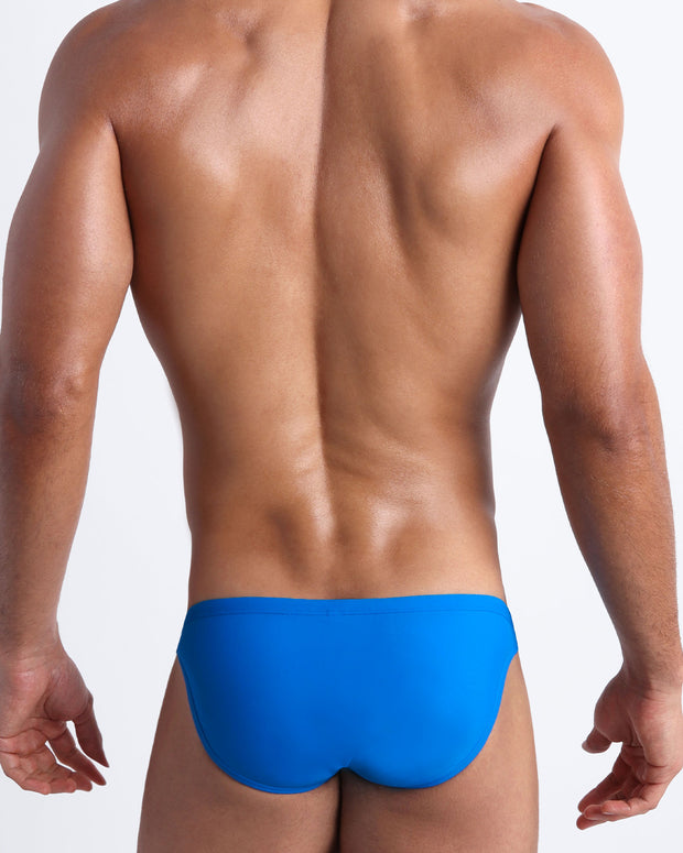 Back view of a male model wearing men’s swim mini-brief in blue by the Bang! Clothes brand of men&