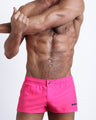 Frontal view of a sexy male model wearing the PINK BOMB men’s square leg swim trunks in a solid hot pink color by the Bang! Menswear brand from Miami.