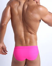 Back view of a male model wearing men’s swim briefs in bright pink color by the Bang! Clothes brand of men's beachwear.