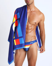 Model showing the STRIPE'A'POSE quick-dry microfiber towel with matching swim briefs for the beach in bright blue with color stripes. 
