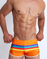 Front view of male model wearing swim shorts in orange color with color stripes in aqua blue, bold red, yellow and dark blue.