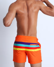 Back view of male model wearing a swim mini-brief in orange color with color stripes in aqua blue, bold red, yellow and dark blue.