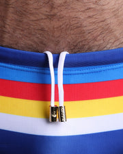 Close-up view of the VIA POSTAL Swim Sunga mens swimsuit in royal blue color with color stripes in white, yellow, bold red, and blue with white internal drawstring cord showing custom branded golden buttons by BANG! clothing brand.
