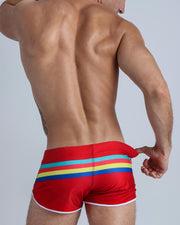Back view of male model wearing a swim mini-brief in red color with color stripes in aqua blue, bold red, yellow and dark blue.