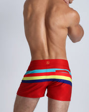 Back side view of male model wearing beach shorts in red color with color stripes in aqua green, bold red, yellow and blue.