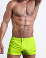 Frontal view of a sexy male model wearing the ULTRA NEON men’s square leg swim trunks in a solid bright lime green color by the Bang! Menswear brand from Miami.