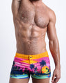 Frontal view of model wearing the UNDER A NEON SKY men’s square leg swim trunks in pink & orange sunset skyline print by the Bang! brand of men's beachwear from Miami.