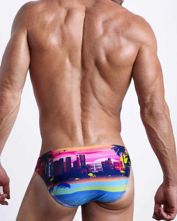 Back view of a model wearing UNDER A NEON SKY men’s beach mini-brief in sunset colors with the miami sunset skyline print made by the Bang! Miami official brand of men&