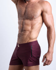 Side view of a masculine model wearing men's VERY BERRY swim summer beach shorts in a wine red  color with official logo of BANG! Brand in gold.